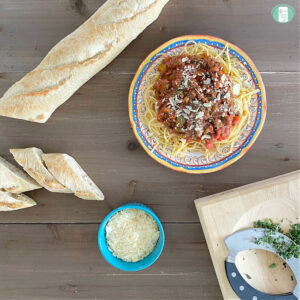 sliced French bread, bowl of grated white cheese, plate of spaghetti, and board with chopped herbs
