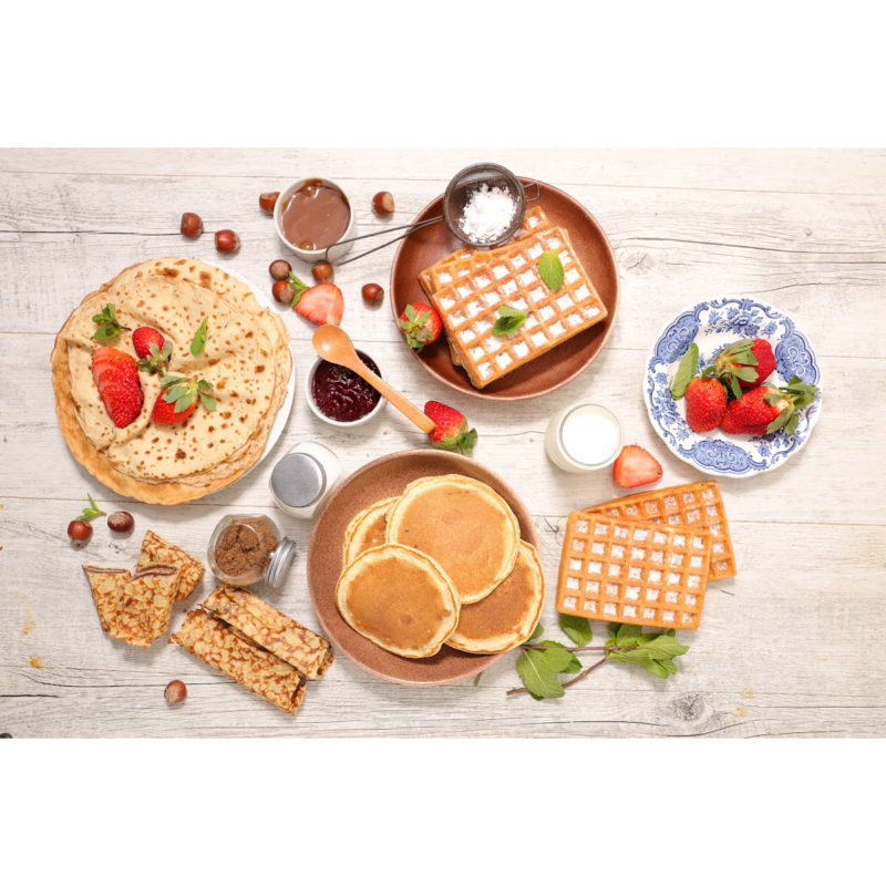 And overhead photo of pancakes, waffes and crepes layed out on a table with syrup, chocolate spread and strawberries.