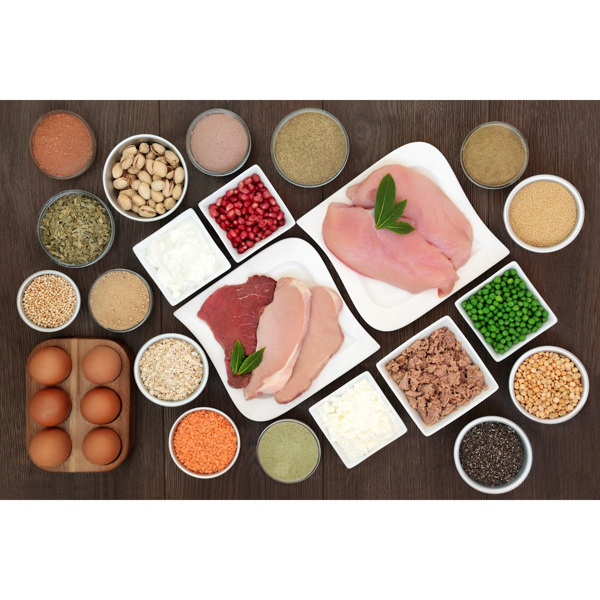 An overhead shot of a variety of foods layed out in a pleasing way. Foods include eggs, raw chicken, raw beef and pork, nuts, tomatoes, peas and other colour foods in bowls.