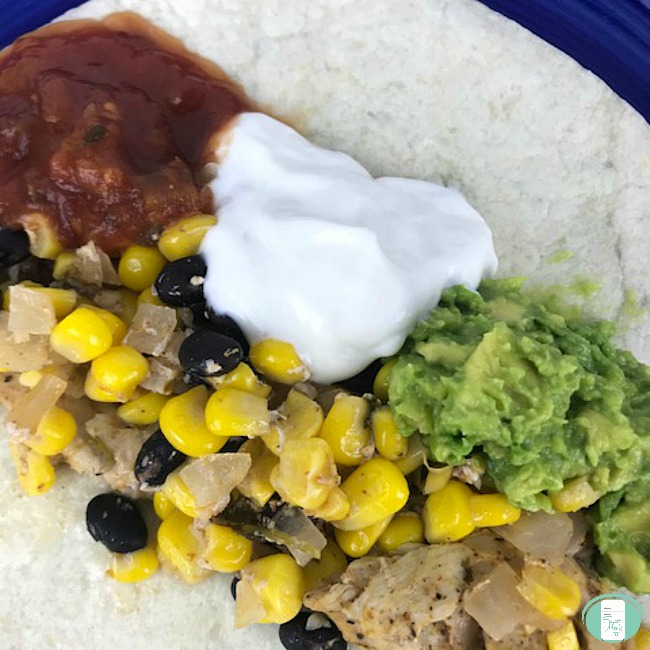 close up of cilantro lime chicken with corn and black beans on a tortilla with guacamole, sour cream and salsa
