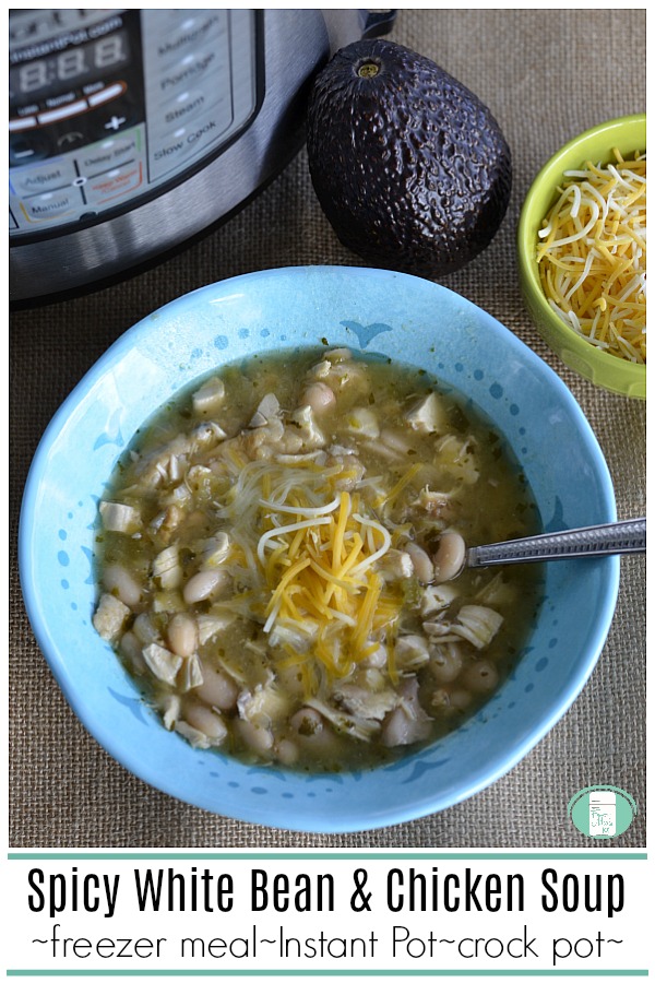a blue bowl filled with white beans, pieces of chicken, topped with shredded cheese and with a spoon in it sits in front of an Instant Pot, an avocado, and a small yellow bowl of shredded cheese. The text reads "Spicy White Bean and Chicken Soup -freezer meal-Instant Pot-crock pot" #freezermeals101 #chickensoup #crockpotmeals #spicywhitebeansoup