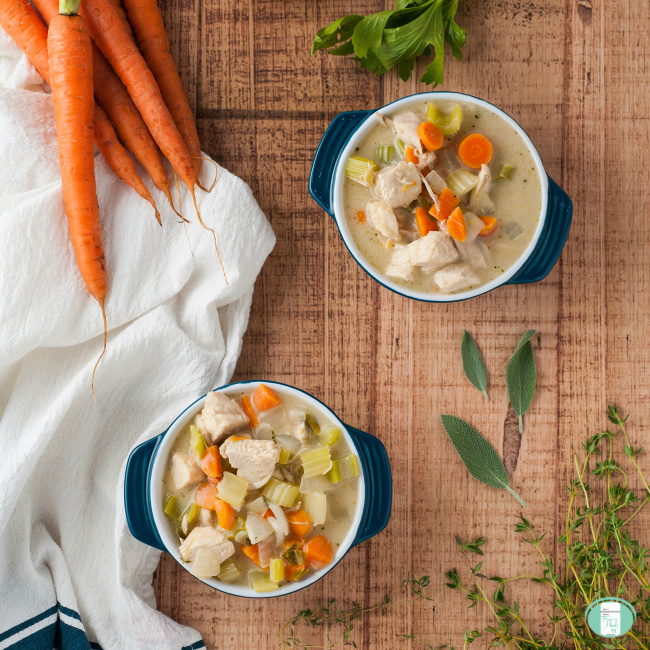 two bowls of chunky cream of chicken soup with a linen napkin, carrots and herbs nearby