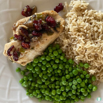 white plate with chicken topped with cranberries next to rice and peas