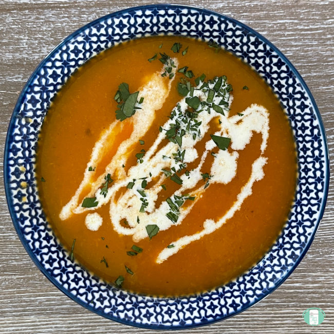 bowl with orange soup topped with drizzles of white cream and sprinkles of green herbs