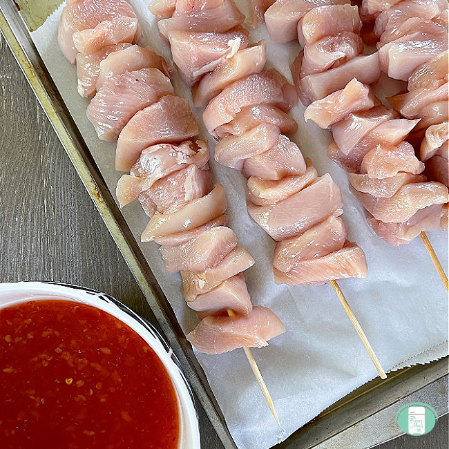 raw chicken cubes on wooden skewers next to a bowl of red sauce