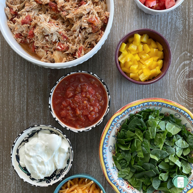 bowls of taco toppings like lettuce, salsa, sour cream, shredded chicken, and cheese