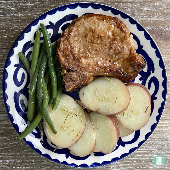 a pork chop, green beans, and sliced cooked potatoes on a blue and white plate