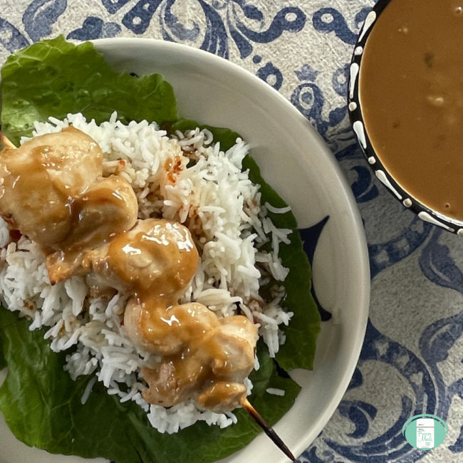 chicken skewers on rice on lettuce next to a bowl of peanut sauce