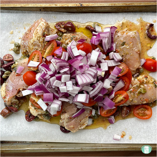 raw chicken with purple onions, olives, baby tomatoes, and seasonings on a baking sheet