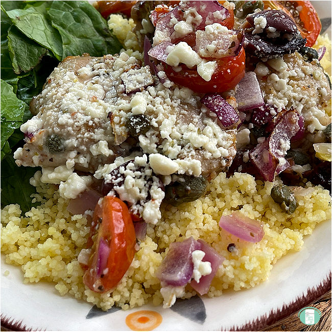 chicken and baby tomatoes and olives topped with feta cheese on top of couscous next to spinach on a plate