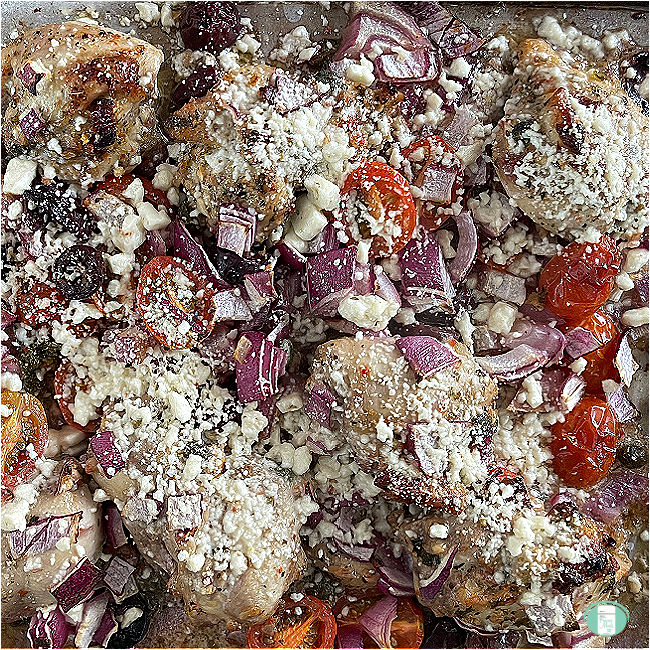 cooked chicken topped with crumbled feta cheese with baby tomatoes and purple onions on a tray