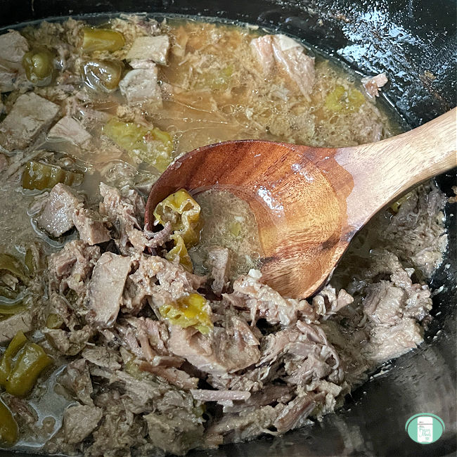 slow cooker with shredded pot roast in gravy in it with a wooden spoon