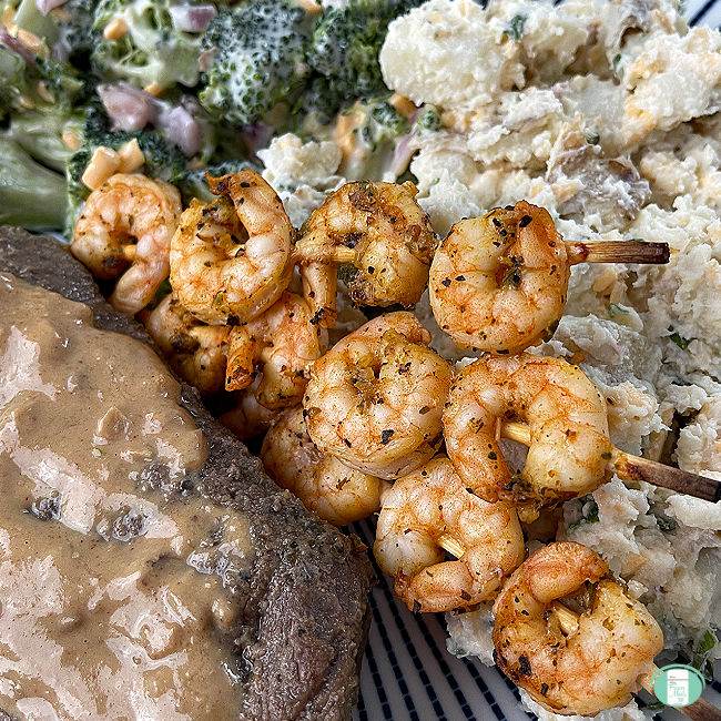 cooked seasoned shrimp on skewers next to steak, potato salad, and broccoli salad