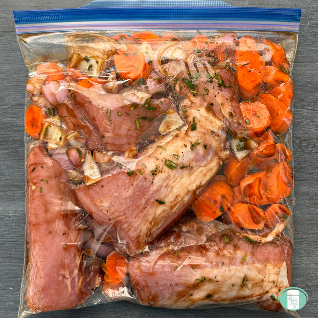 Close up of a freezer quality bag with pork tenderloin, seasonings, onion, and chopped carrots.