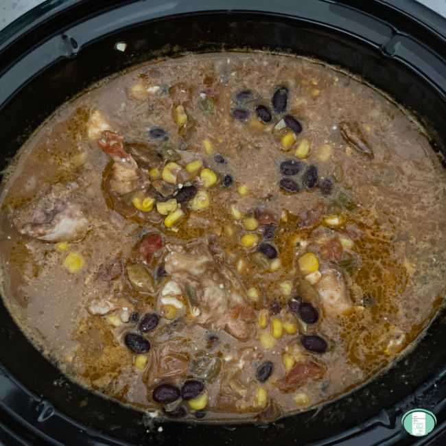 Tex Mex Chicken in a crock pot with the lid off. Bits of chicken along with a sprinkle of kernel corn and black beans are floating on top of the rich brown liquid in the pot.