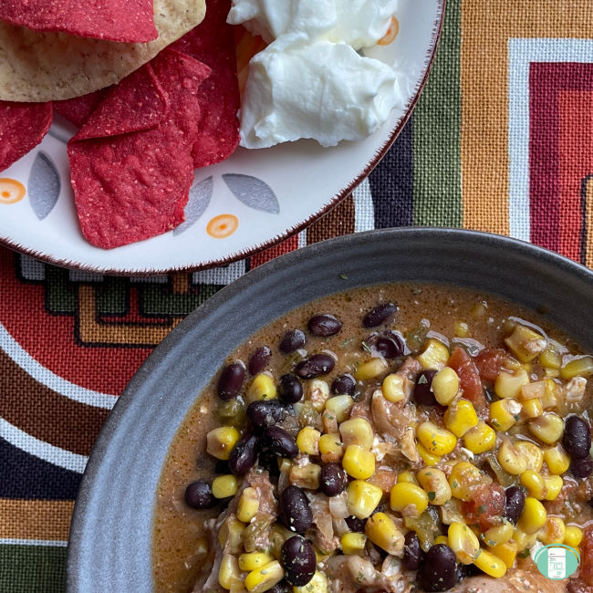 A bowl of cooked Tex Mex chicken with kernel corn and black beans beside a plate with some festive taco chips and a blob of sour cream