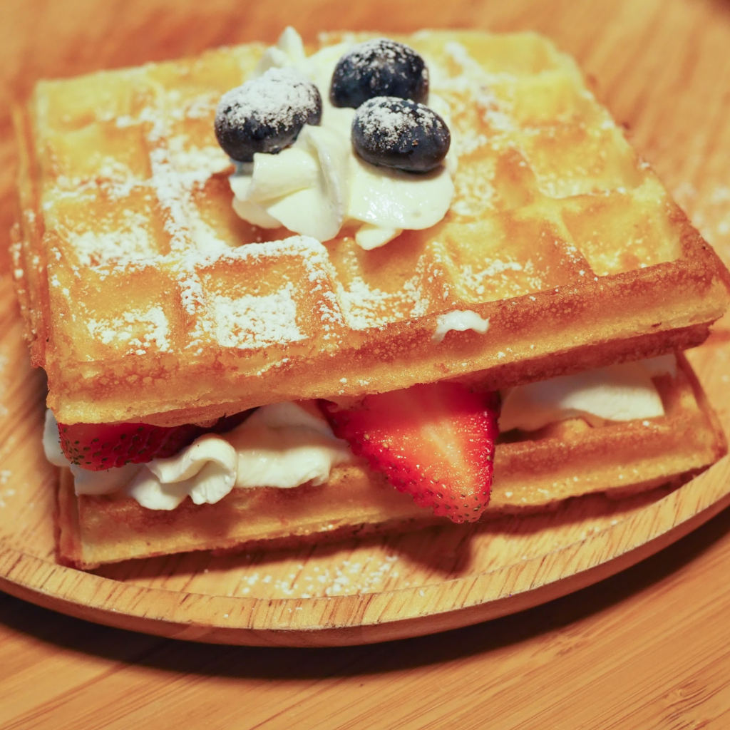 Buttermilk Waffles stacked with whipped cream and strawberries sandwiched in between, and topped with more whipped cream and blueberries and a dusting of icing sugar