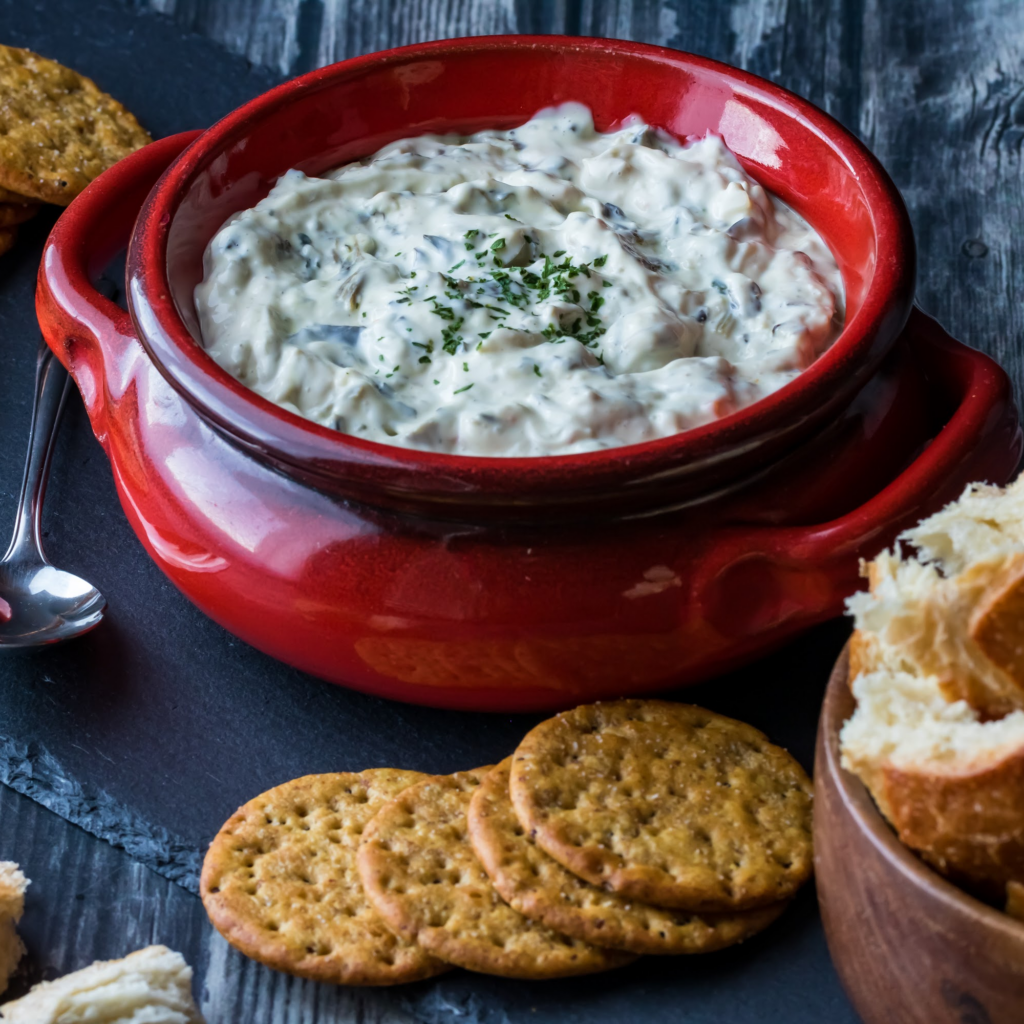 Spinach and Artichoke Dip served in a festive red casserole dish, accompanied with crackers a cubes of sour dough bread