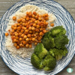Buffalo Chickpeas served over rice with broccoli on the side, on a striped blue and white plate sitting on a wooden table. Top down image showing the whole plate.