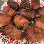 Close-up image of cooked bacon-wrapped blue cheese stuffed dates arranged on a festive plate. The bacon is crispy and golden brown, while the dates appear glossy and caramelized, with some cheese slightly visible at the ends.