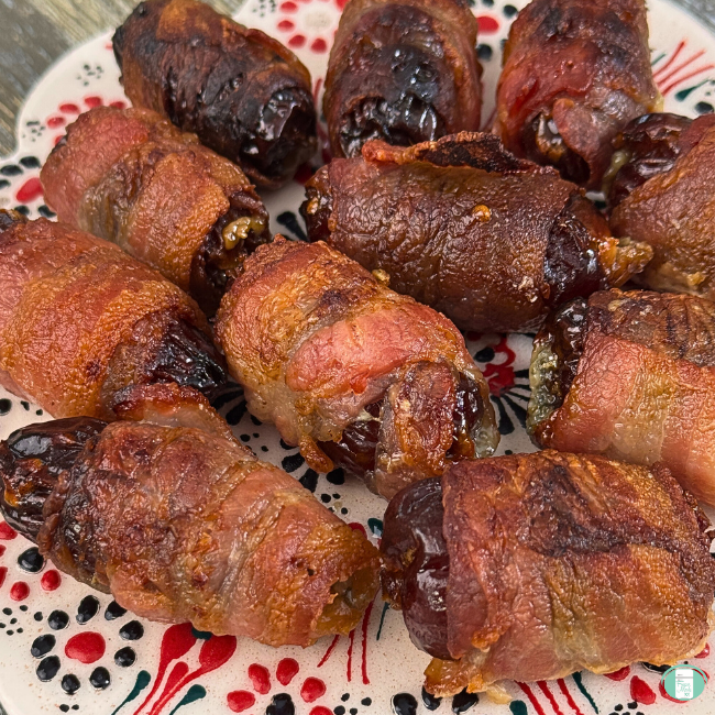 Close-up image of cooked bacon-wrapped blue cheese stuffed dates arranged on a festive plate. The bacon is crispy and golden brown, while the dates appear glossy and caramelized, with some cheese slightly visible at the ends.