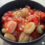 Close-up of Gnocchi al’Arrabbiata in a black bowl, featuring tender potato gnocchi coated in a chunky tomato and red pepper sauce with bits of minced onion and sprinkled herbs.