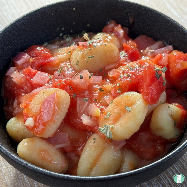 Close-up of Gnocchi al’Arrabbiata in a black bowl, featuring tender potato gnocchi coated in a chunky tomato and red pepper sauce with bits of minced onion and sprinkled herbs.