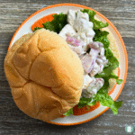 Lemon Pepper Chicken Bites on a bun over lettuce and tomato. Served on a white plate with orange and yellow rim resting on a grey background.