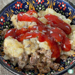 Serving of tater tot casserole with ground sausage, topped with ketchup, in a decorative black bowl with colorful floral patterns.