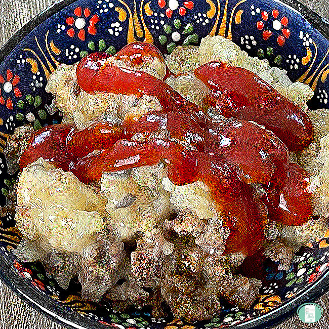 Serving of tater tot casserole with ground sausage, topped with ketchup, in a decorative black bowl with colorful floral patterns.