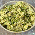 Close-up of a bowl filled with Green Pasta Salad made with bow tie pasta, green peas, chopped artichoke hearts, and a creamy pesto and ranch-style dressing. The pasta and peas are coated evenly, and the bowl sits on a light wood surface.