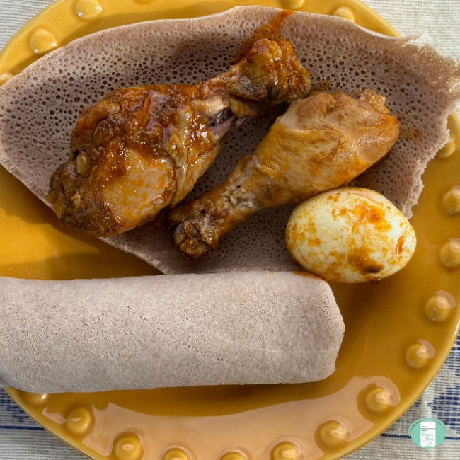 Plated Doro Wat with two chicken legs and a boiled egg covered in sauce served over injera with a bumpy yellow plate.