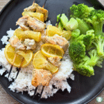 Marinated salmon skewers with lemon slices served over white rice alongside steamed broccoli on a black plate.