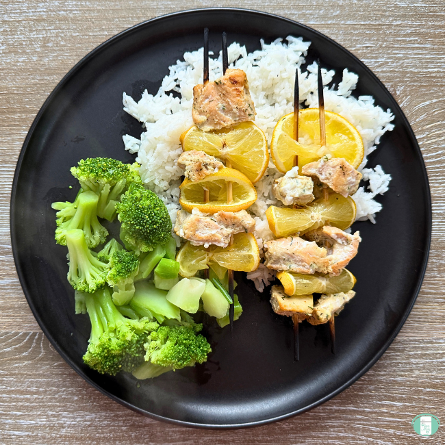 Marinated salmon skewers with lemon slices served over white rice with steamed broccoli on a black plate, photographed from above.