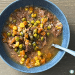 Bowl of Texas Beef Soup filled with tender beef, peas, corn, carrots, and onions in a savory broth, garnished with herbs and served with a spoon.