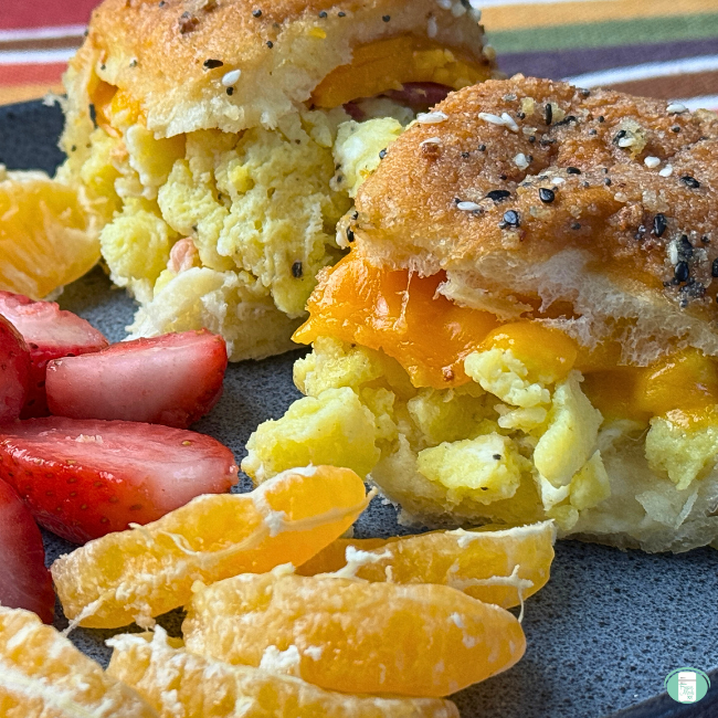Close-up of two breakfast sliders with everything bagel–seasoned buns, filled with fluffy scrambled eggs, melted cheddar, and bacon, served on a plate with sliced strawberries and orange segments.