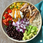 Mexican marinated chicken bowl with sliced grilled chicken, white rice, black beans, corn, diced tomatoes, red onion, avocado, sour cream, and a drizzle of orange sauce.
