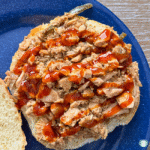 Open-faced Texas pulled pork sandwich topped with tender shredded pork, sliced jalapeños, and barbecue sauce on a soft bun, served on a blue plate.
