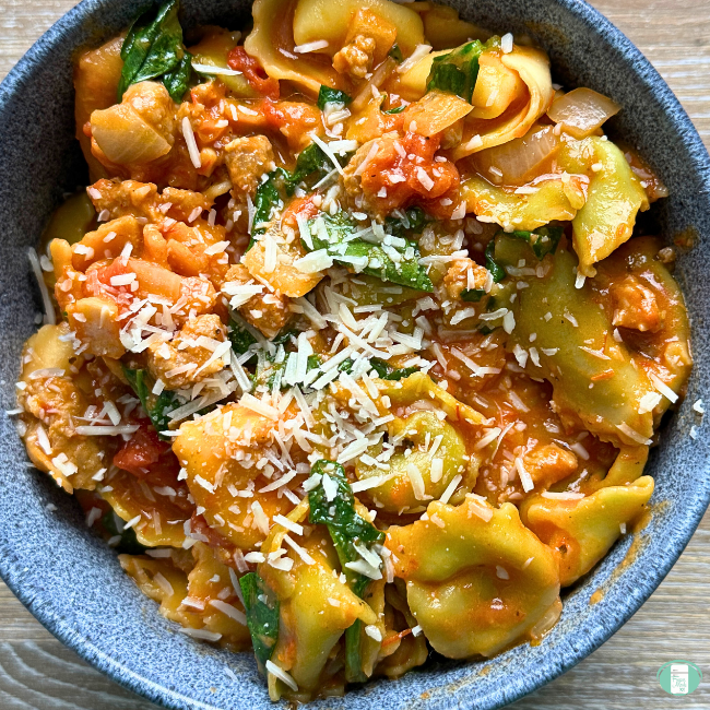 blue bowl filled with tortellini, sausage, and spinach topped with Parmesan cheese