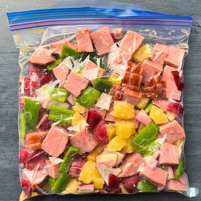 Chunks of ham, green pepper, purple onion and pineapple in clear bag.