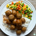 Honey bourbon meatballs served on a white plate over rice with a side of frozen vegetables.