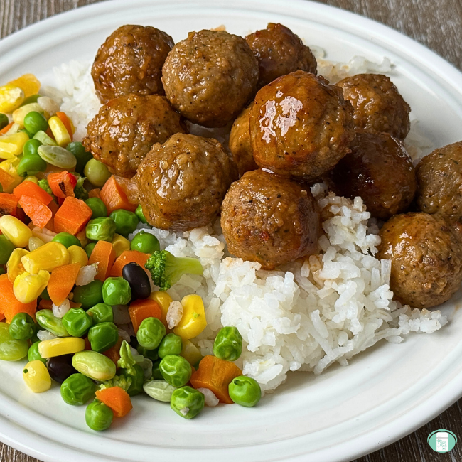 Honey bourbon meatballs served on a white plate over rice with a side of frozen vegetables.