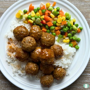 Honey bourbon meatballs served on a white plate over rice with a side of frozen vegetables.