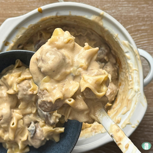 Creamy stroganoff sauce over meatballs and noodles in a serving spoon, held overtop of a crock pot and a bowl of the meal