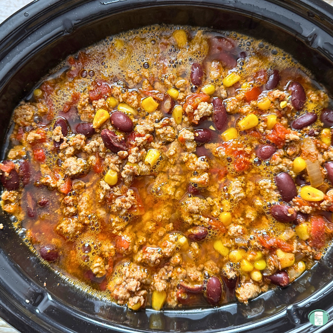 Turkey chili cooking in a crock pot with ground turkey, beans, onions, corn, and seasonings