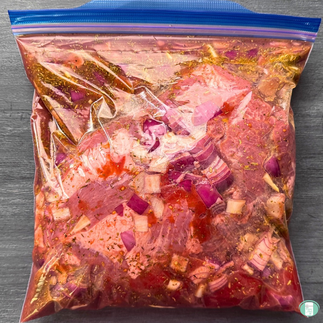 Freezer meal pork roast ragu in a ziploc bag with pork shoulder, red onion, and a colourful, well seasoned sauce, ready to freeze