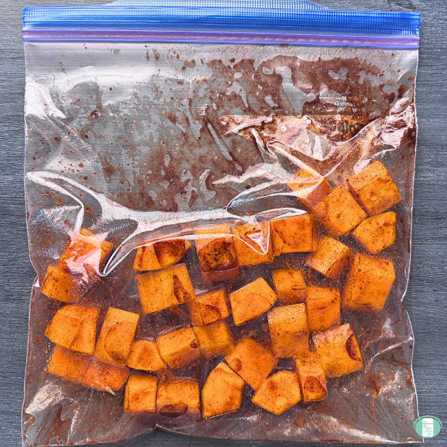 Cubed butternut squash covered in cinnamon, nutmeg, maple syrup, and other seasonings in ziploc bag, ready to go in the freezer