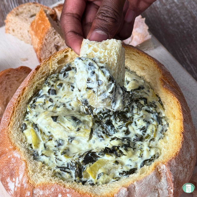 Spinach artichoke dip in a bread bowl, with a small chunk of bread dipped in it